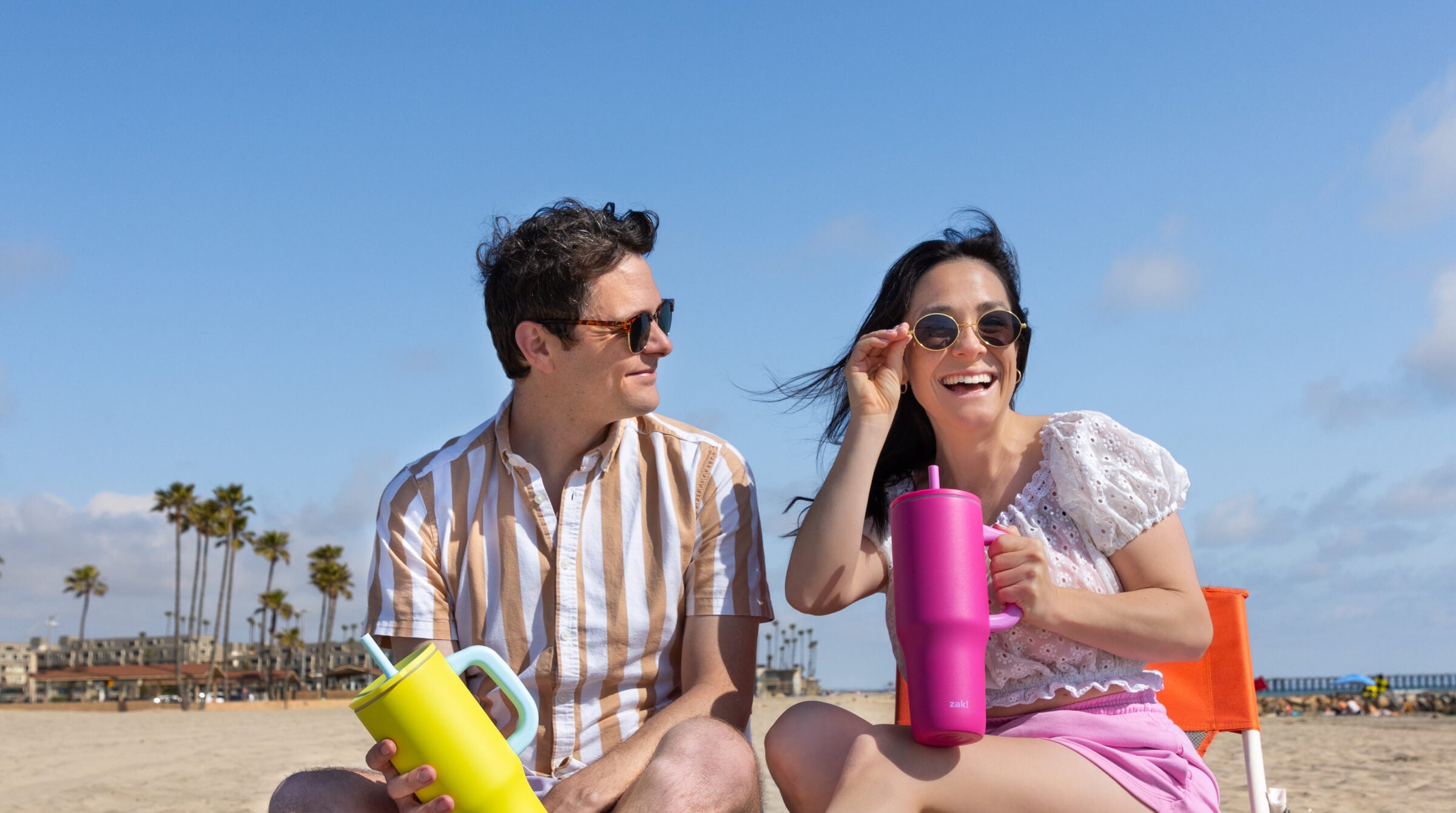 Two people sitting on a beach with colorful zak cups, palm trees in the background